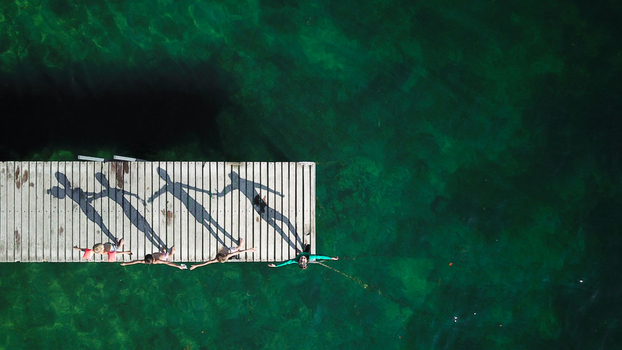Aerial view of four children standing on a wooden pier with arms outstretched casting long shadows over turquoise water in Saint Prex Vaud Switzerland.