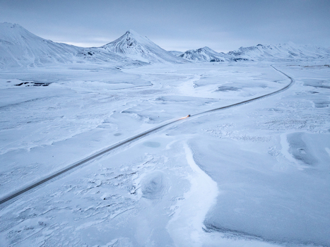 Aerial view of a lonely road winding through a vast snow covered landscape with distant mountains under a soft sky in Egilsstadir Mulathing Iceland. Aerial view of a lonely road winding through a vast snow covered landscape with distant mountains under a soft sky in Egilsstadir Mulathing Iceland.