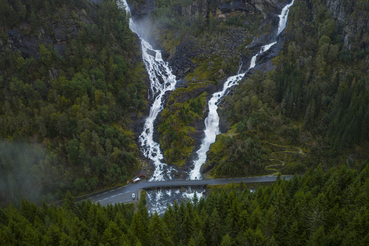 Aerial view of Latefossen twin waterfalls cascading down rocky cliffs through a dense pine forest with a road bridge at the bottom Skare Vestland Norway.