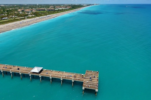 Aerial view of the Juno Beach Pier stretching over turquoise Atlantic waters toward a sandy coastline with beachgoers and coastal buildings Juno Beach Florida United States. Aerial view of the Juno Beach Pier stretching over turquoise Atlantic waters toward a sandy coastline with beachgoers and coastal buildings Juno Beach Florida United States.
