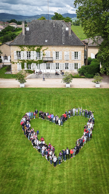 La Chapelle de Guinchay France 01 June 2024 Aerial view of a large group of people forming a heart shape on a green lawn in front of a historic stone manor house.
