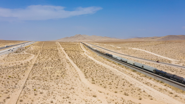 Aerial view of a long freight train traveling through a vast desert landscape alongside a highway under a clear blue sky in the United States.