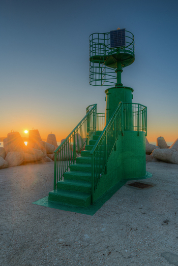 View of a green beacon with metal stairs on a breakwater surrounded by concrete tetrapods during a golden sunrise in Fano Marche Italy. View of a green beacon with metal stairs on a breakwater surrounded by concrete tetrapods during a golden sunrise in Fano Marche Italy.