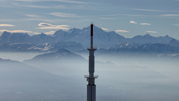 Aerial view of a tall tower piercing through the fog with snow capped mountains rising majestically in the background Gex Auvergne Rhône Alpes France. Aerial view of a tall tower piercing through the fog with snow capped mountains rising majestically in the background Gex Auvergne Rhône Alpes France.