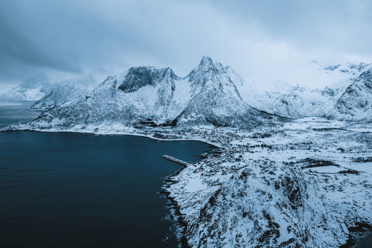 Aerial view of the stark snow laden peaks rising majestically above the dark waters a tranquil fjord embracing the quiet village nestled below Mefjordvær Troms Norway. Aerial view of the stark snow laden peaks rising majestically above the dark waters a tranquil fjord embracing the quiet village nestled below Mefjordvær Troms Norway.