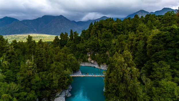 Aerial view of a suspension bridge stretching over the turquoise waters embraced by dense green forests and framed by distant hazy mountains Hokitika Gorge New Zealand. Aerial view of a suspension bridge stretching over the turquoise waters embraced by dense green forests and framed by distant hazy mountains Hokitika Gorge New Zealand.