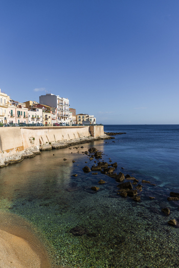 View of the clear turquoise sea gently caressing the rocky shore beneath the ancient stone walls of the city s edge Ortigia Sicily Italy.