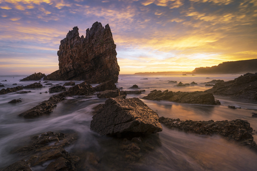 View of the iconic sea stack at Playa de Portizuelo with misty water and a golden sunset sky over the rocky coastline Spain. View of the iconic sea stack at Playa de Portizuelo with misty water and a golden sunset sky over the rocky coastline Spain.