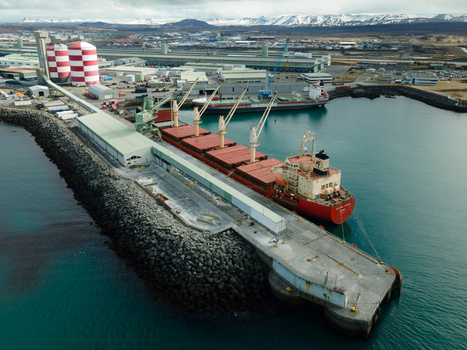 Hafnarfjordur Iceland 17 April 2026 Aerial view of the industrial port with a large cargo ship docked alongside a conveyor system and red striped silos against a backdrop of snow capped mountains. Hafnarfjordur Iceland 17 April 2026 Aerial view of the industrial port with a large cargo ship docked alongside a conveyor system and red striped silos against a backdrop of snow capped mountains.
