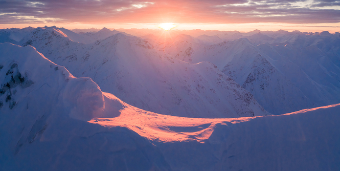 Aerial view of snow capped mountain peaks bathed in the warm glow of the setting sun creating a stunning contrast against the cool blue shadows Anaktuvuk Pass Alaska United States.