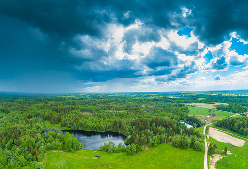 Aerial view of a serene lake nestled within a lush green forest and rolling meadows under a dramatic sky with heavy storm clouds in Latvia. Aerial view of a serene lake nestled within a lush green forest and rolling meadows under a dramatic sky with heavy storm clouds in Latvia.