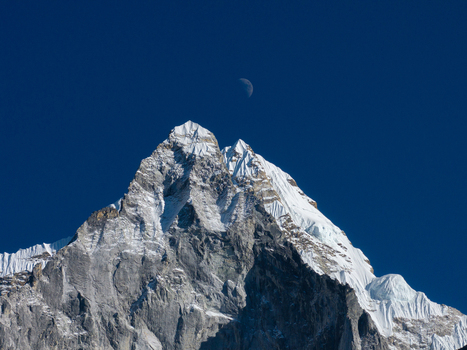 Aerial view of a snow capped Himalayan mountain peak with jagged rock formations and a crescent moon visible in the clear deep blue sky in Nepal.