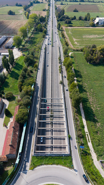Aerial view of a provincial road featuring a partially covered tunnel section with cars driving through surrounded by green agricultural fields and trees in Cremona Lombardia Italy.