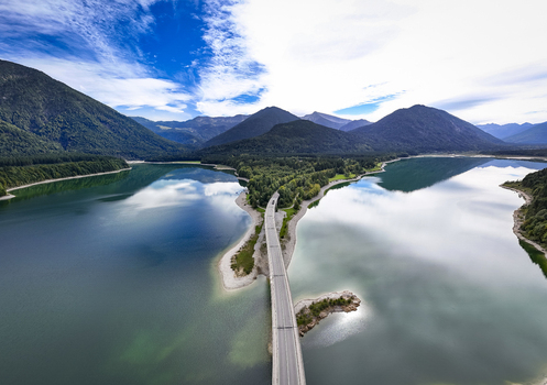 Aerial view of a long bridge cutting across the tranquil waters of Sylvensteinsee reflecting the vibrant sky and towering mountains Sylvensteinspeicher Bayern Germany. Aerial view of a long bridge cutting across the tranquil waters of Sylvensteinsee reflecting the vibrant sky and towering mountains Sylvensteinspeicher Bayern Germany.