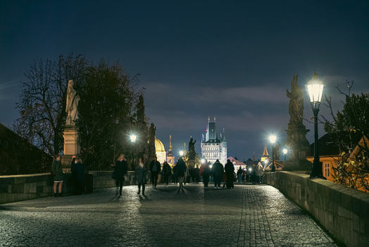 Prague Czechia 03 November 2021 View of Charles Bridge at night with illuminated street lamps stone statues and people walking along the cobblestone path towards the bridge tower. Prague Czechia 03 November 2021 View of Charles Bridge at night with illuminated street lamps stone statues and people walking along the cobblestone path towards the bridge tower.