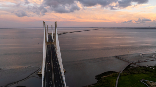 Aerial view of Vasco da Gama Bridge stretching across the Tagus River under a dramatic sunset sky with soft clouds Lisboa Portugal.