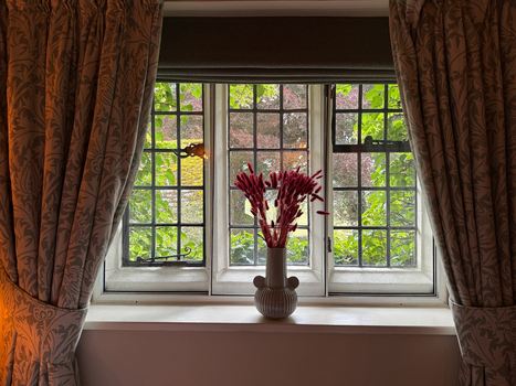 View of a leaded glass window with a vase of red flowers on the sill and patterned curtains looking out onto a lush green garden in Hawes England United Kingdom. View of a leaded glass window with a vase of red flowers on the sill and patterned curtains looking out onto a lush green garden in Hawes England United Kingdom.