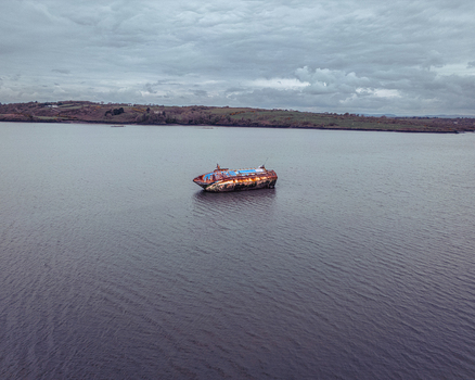 Aerial view of a rusty abandoned shipwreck floating in the calm waters of a wide river with green hills along the shoreline under an overcast sky. Aerial view of a rusty abandoned shipwreck floating in the calm waters of a wide river with green hills along the shoreline under an overcast sky.