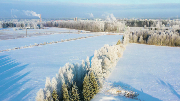 Aerial view of a frost covered winter landscape with snow covered fields lines of rime covered trees and industrial chimneys emitting smoke in the distance in Latvia. Aerial view of a frost covered winter landscape with snow covered fields lines of rime covered trees and industrial chimneys emitting smoke in the distance in Latvia.