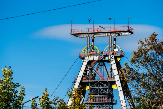 View of a historic mine headframe with yellow winding wheels and steel structure surrounded by autumn trees under a clear blue sky in Poland.