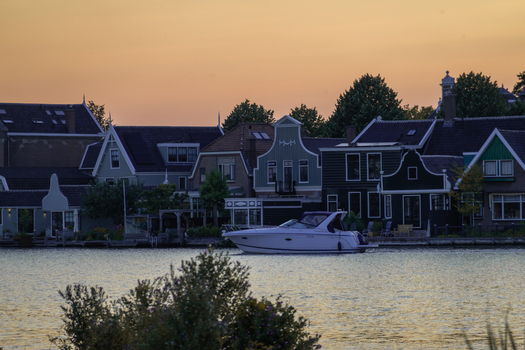 View of a motorboat cruising on the Zaan river in front of traditional wooden houses at Zaanse Schans during a warm sunset in Koog aan de Zaan North Holland Netherlands.