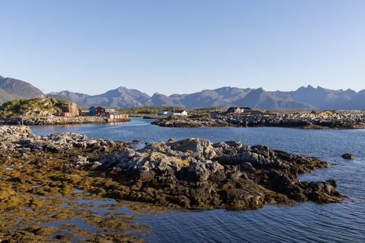 View of the rocky coastline and traditional fishing village houses under a clear blue sky with mountains in the background Laukvik Nordland Norway. View of the rocky coastline and traditional fishing village houses under a clear blue sky with mountains in the background Laukvik Nordland Norway.