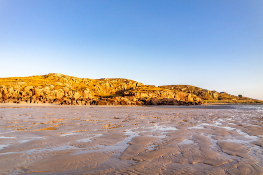 View of the sandy beach at low tide with rocky golden hills under a clear blue sky in Cloughglass County Donegal Ireland. View of the sandy beach at low tide with rocky golden hills under a clear blue sky in Cloughglass County Donegal Ireland.