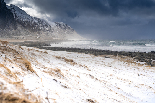 View of a snow covered beach with rocky coastline and dramatic dark clouds over the mountains in Leknes Lofoten Norway.
