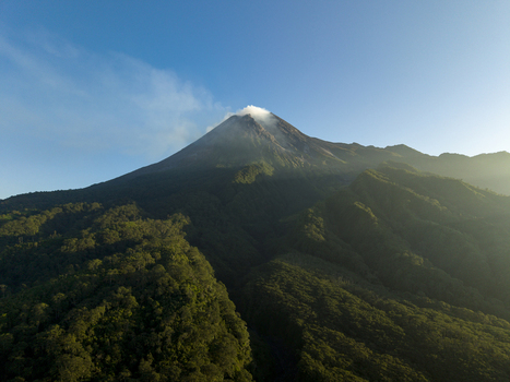 Aerial view of Mount Merapi with smoke rising from the peak and lush green forests on the slopes in Kecamatan Pakem Daerah Istimewa Yogya Indonesia. Aerial view of Mount Merapi with smoke rising from the peak and lush green forests on the slopes in Kecamatan Pakem Daerah Istimewa Yogya Indonesia.