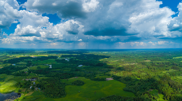 Aerial view of lush green forests and fields with small ponds under a dramatic sky with heavy clouds and patches of sunlight in Latvia. Aerial view of lush green forests and fields with small ponds under a dramatic sky with heavy clouds and patches of sunlight in Latvia.
