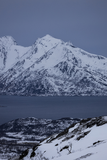 View of snow capped mountain peaks rising above a calm fjord under a moody blue winter sky Kvaloya Troms Norway.