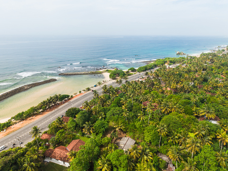 Aerial view of the tropical coastline with a coastal road lush palm trees and turquoise ocean waves in Koggala Southern Province Sri Lanka. Aerial view of the tropical coastline with a coastal road lush palm trees and turquoise ocean waves in Koggala Southern Province Sri Lanka.