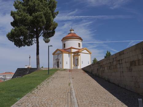 View of the Calvary Chapel with its white walls and orange roof situated next to a stone wall and a tall tree along a cobblestone path in Portalegre Portalegre Portugal. View of the Calvary Chapel with its white walls and orange roof situated next to a stone wall and a tall tree along a cobblestone path in Portalegre Portalegre Portugal.