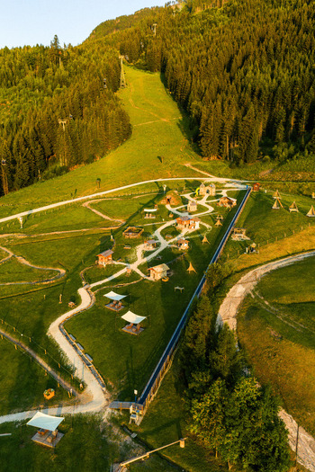 Aerial view of Fiemme Valley featuring lush green meadows winding paths small wooden structures and dense pine forests under warm golden hour light in Trentino Alto Adige Italy.