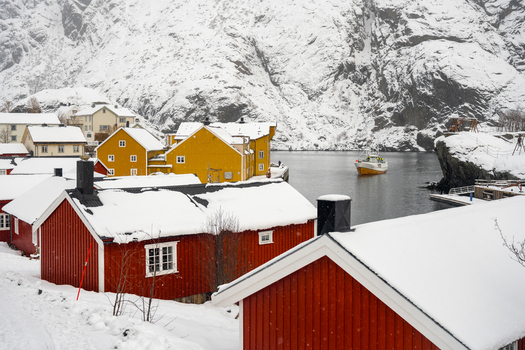 View of traditional red and yellow rorbuer cabins covered in snow along the icy coastline with a fishing boat in the water in Leknes Lofoten Norway.