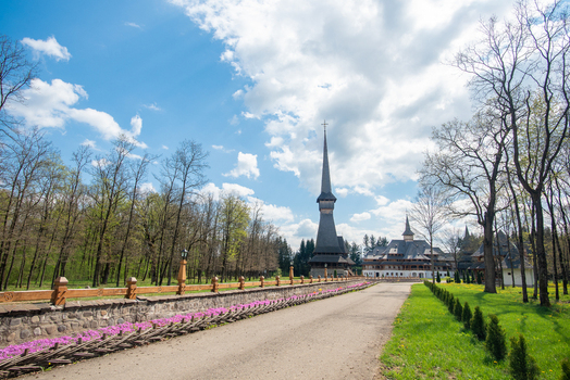 View of the traditional wooden church with a tall spire in the Maramures region surrounded by lush trees a stone wall and a gravel path in Maramures Romania. View of the traditional wooden church with a tall spire in the Maramures region surrounded by lush trees a stone wall and a gravel path in Maramures Romania.