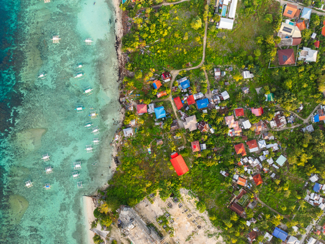 Aerial view of the turquoise coastline of Panagsama Beach with outrigger boats and colorful rooftops nestled in tropical greenery in Moalboal Central Visayas Philippines.