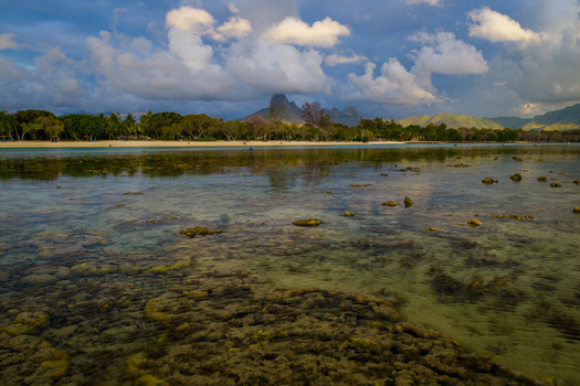 Aerial view of the shallow coral reefs and sandy beach with lush tropical trees and mountains under a cloudy sky in Moka District Mauritius. Aerial view of the shallow coral reefs and sandy beach with lush tropical trees and mountains under a cloudy sky in Moka District Mauritius.