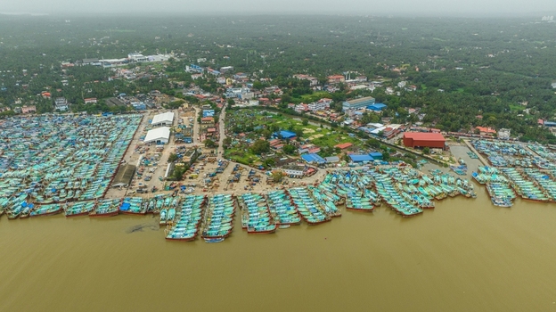 Aerial view of Malpe Fishing Harbour with numerous colorful fishing boats docked along the muddy riverbank surrounded by lush green vegetation and urban structures in Udupi Karnataka India. Aerial view of Malpe Fishing Harbour with numerous colorful fishing boats docked along the muddy riverbank surrounded by lush green vegetation and urban structures in Udupi Karnataka India.