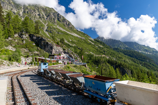 View of the VerticAlp Emosson mountain railway with vintage mining wagons on tracks set against lush alpine slopes rocky cliffs and blue sky in Le Chatelard Fribourg Switzerland. View of the VerticAlp Emosson mountain railway with vintage mining wagons on tracks set against lush alpine slopes rocky cliffs and blue sky in Le Chatelard Fribourg Switzerland.