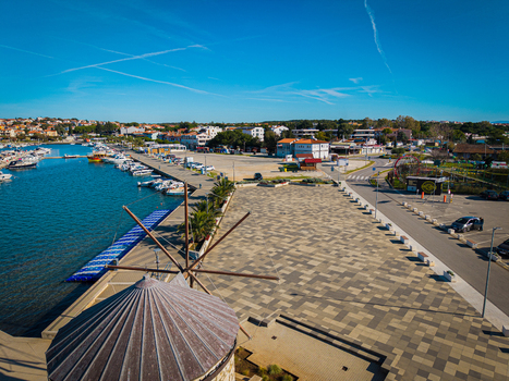 Aerial view of a stone paved square contrasts with the azure harbor dotted with boats overlooked by terracotta rooftops under a clear sky Medulin Istria County Croatia. Aerial view of a stone paved square contrasts with the azure harbor dotted with boats overlooked by terracotta rooftops under a clear sky Medulin Istria County Croatia.