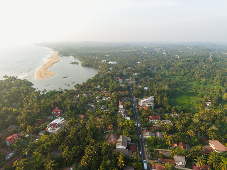 Aerial view of the tropical coastline with a sandy beach a lagoon and a straight road cutting through dense palm trees and residential houses in Galle Southern Province Sri Lanka.