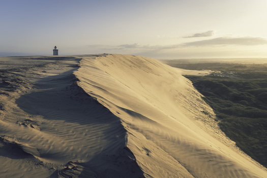 View of the Rubjerg Knude lighthouse standing on the edge of a massive sand dune at sunset with soft golden light and coastal vegetation in Lokken Denmark. View of the Rubjerg Knude lighthouse standing on the edge of a massive sand dune at sunset with soft golden light and coastal vegetation in Lokken Denmark.