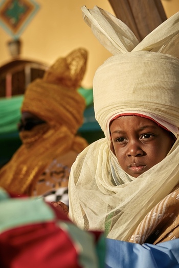 Zaria City Nigeria 21 March 2026 View of a child in regal cream colored traditional attire with a solemn gaze bathed in warm golden light amidst vibrant fabrics. Zaria City Nigeria 21 March 2026 View of a child in regal cream colored traditional attire with a solemn gaze bathed in warm golden light amidst vibrant fabrics.