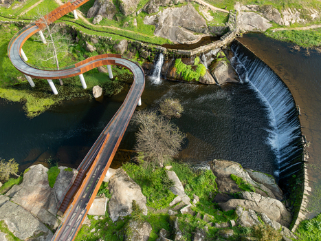 Aerial view of Acude da Ribeira featuring a modern winding walkway over a river dam with waterfalls and rocky terrain in Ervedal Coimbra Portugal. Aerial view of Acude da Ribeira featuring a modern winding walkway over a river dam with waterfalls and rocky terrain in Ervedal Coimbra Portugal.