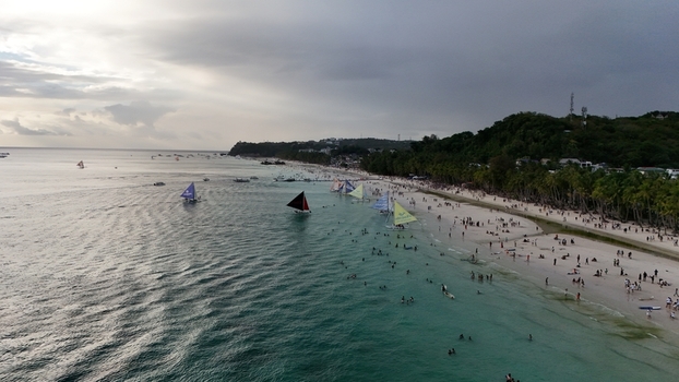 Malay Philippines 02 May 2026 Aerial view of Boracay Island featuring sailboats on turquoise water and people on the white sand beach lined with palm trees under a cloudy sky.