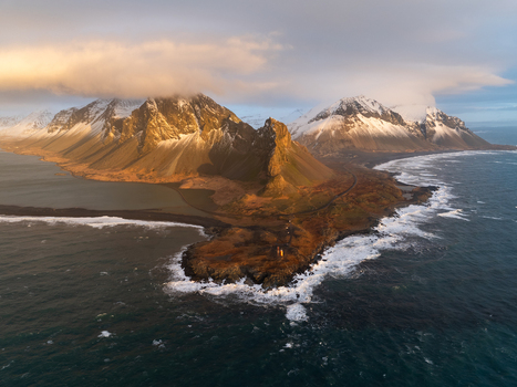 Aerial view of Hvalnes Lighthouse situated on a rocky coastline with dramatic snow capped mountains in the background under golden hour light in Sveitarfelagid Hornafjordur Iceland.
