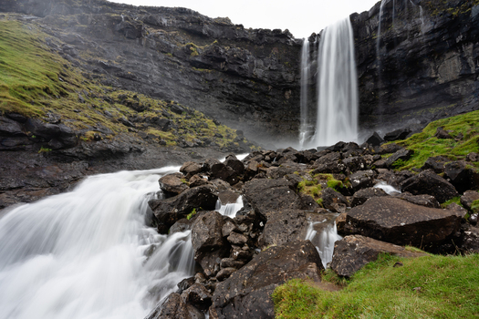 View of a powerful waterfall cascading over dark volcanic cliffs into a rocky stream bed surrounded by lush green mossy slopes in Faroe Islands.