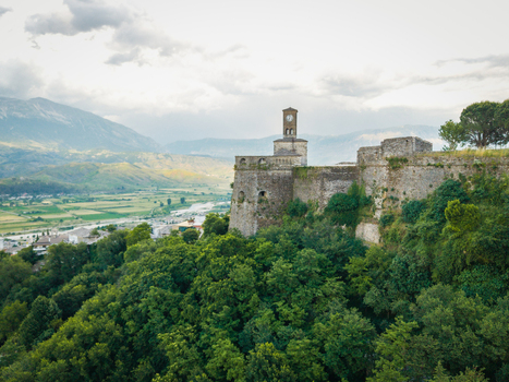 Aerial view of the historic Gjirokaster Fortress with its clock tower overlooking a lush valley and mountains under a cloudy sky Gjirokaster Gjirokaster County Albania. Aerial view of the historic Gjirokaster Fortress with its clock tower overlooking a lush valley and mountains under a cloudy sky Gjirokaster Gjirokaster County Albania.