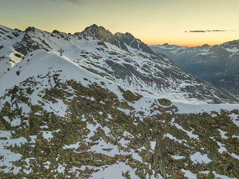 Aerial view of Vallespluga mountain peak featuring a summit cross set against a backdrop of snow covered slopes and golden hour sky in Lombardy Italy. Aerial view of Vallespluga mountain peak featuring a summit cross set against a backdrop of snow covered slopes and golden hour sky in Lombardy Italy.
